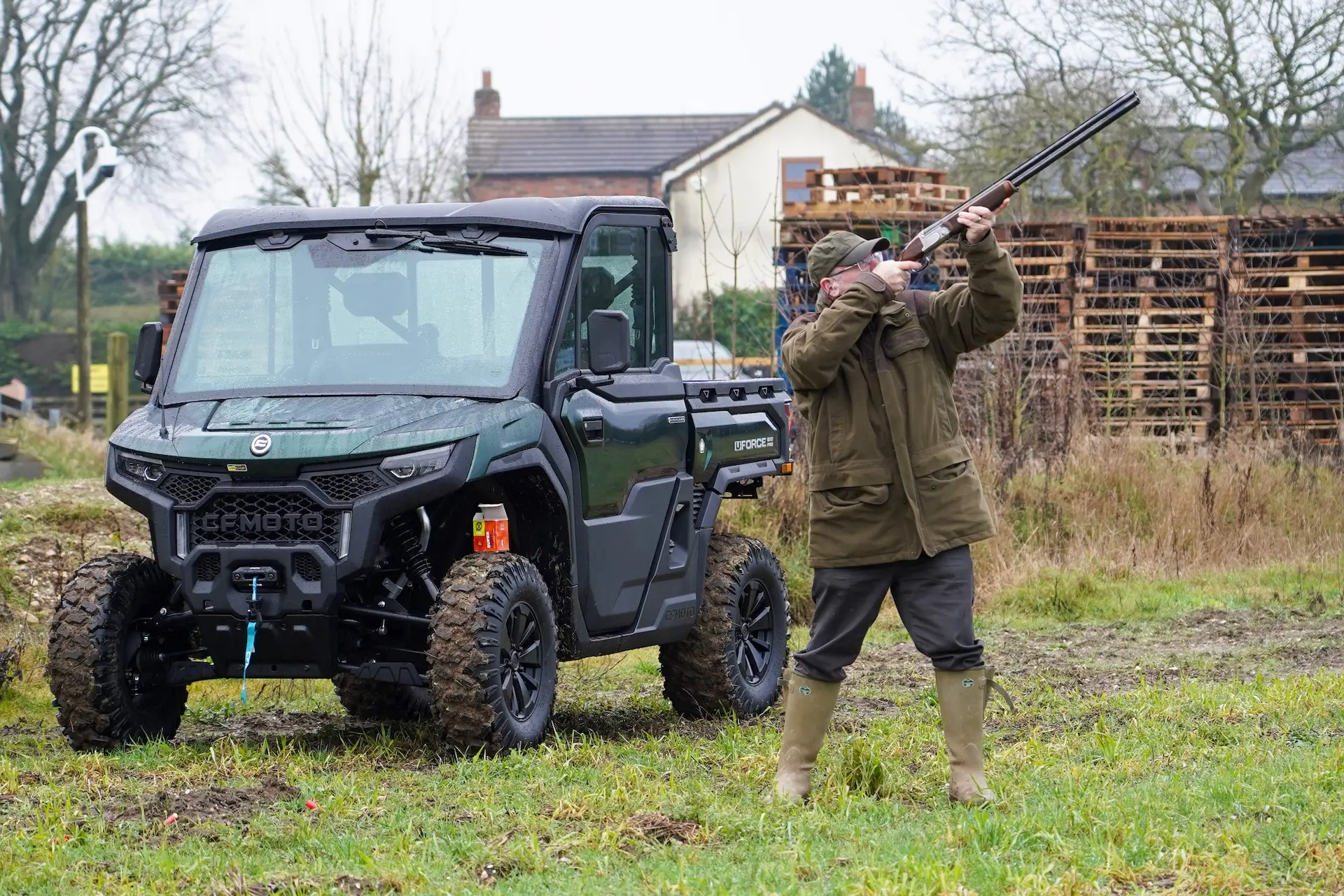 A person in outdoor clothing aims a shotgun upwards beside an off-road utility vehicle, raising the question—what are OTR charges? The rural setting features grass, pallets, and houses in the background.