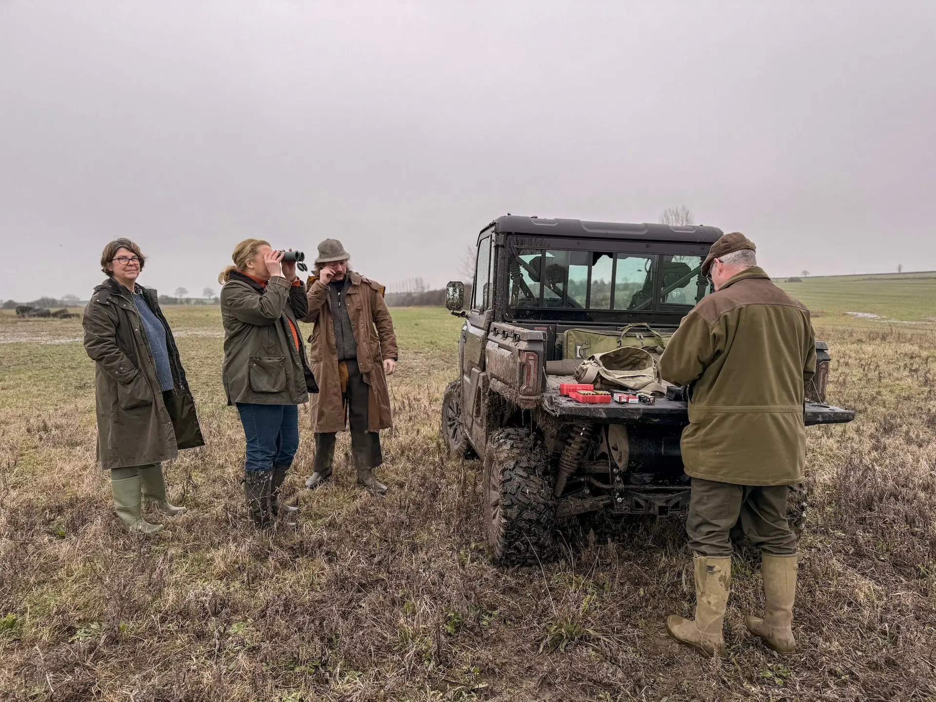 Four people stand beside an off-road vehicle in a field; one looks through binoculars, another writes on a pad, and two others discuss what OTR charges are as they watch the scene unfold.