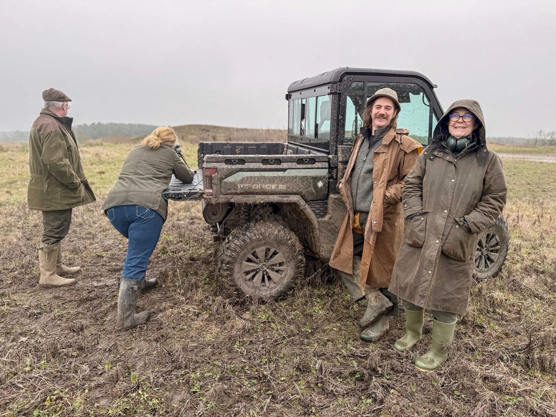Four people in outdoor clothing stand by a muddy utility vehicle in a grassy field on a cloudy day. As they inspect the vehicle’s bed, a sign nearby poses the question: What are OTR charges?.