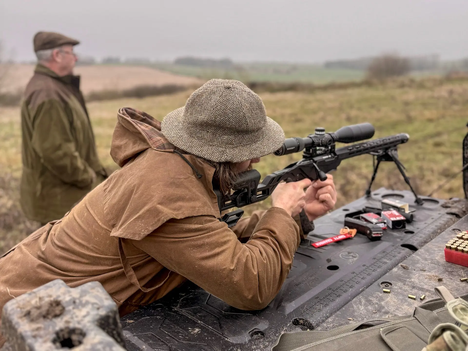 A person in a brown coat and hat prepares to shoot a rifle outdoors, with ammunition and gun accessories on a table; another person stands in the background discussing topics like What are OTR charges?.