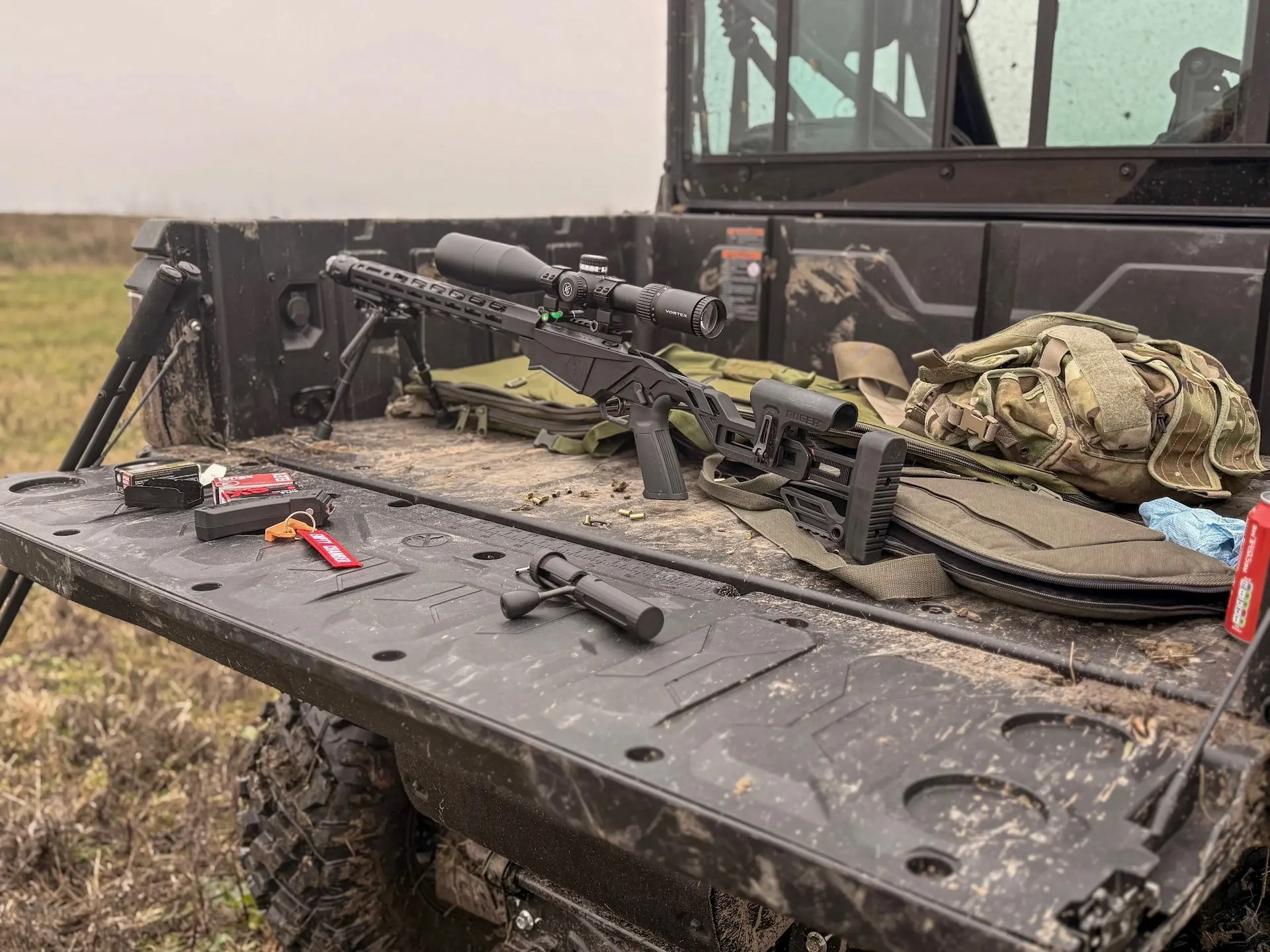 A rifle with a scope, ammunition, a suppressor, and a rucksack are laid out on the tailgate of a muddy utility vehicle in an outdoor setting—much like preparing for an expedition or asking, what are OTR charges before setting off.