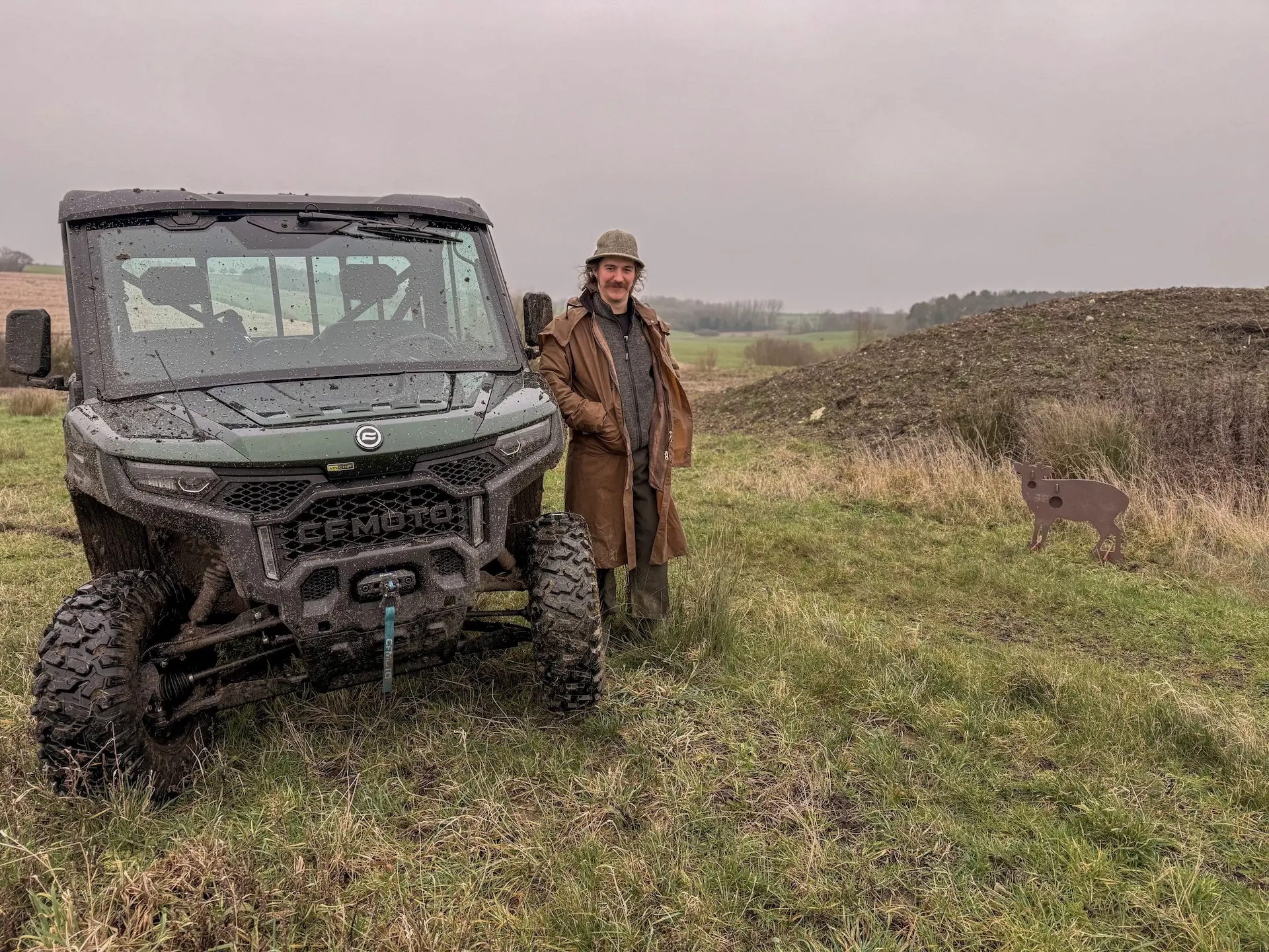A person in a brown coat and hat stands beside a muddy off-road vehicle on grass, with a small deer target nearby under a cloudy sky—leaving you to wonder, what are OTR charges for adventures like this?.