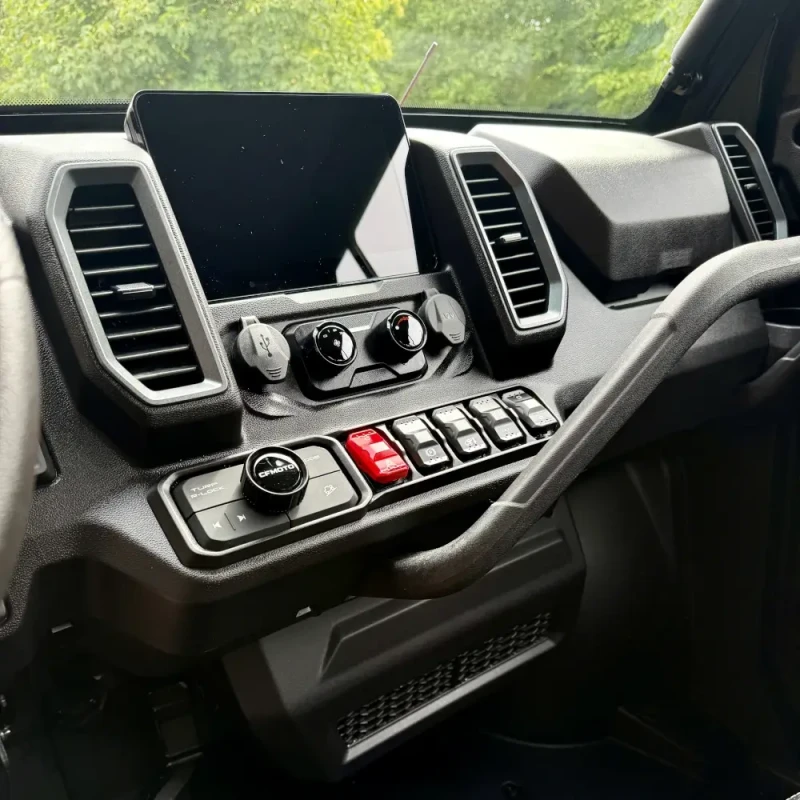 Dashboard of the UFORCE U10 PRO HIGHLAND with a central display, control knobs, buttons, and switches, viewed from the passenger side, with trees visible through the windshield.