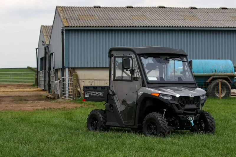 CFMOTO UTV maintenance tips A black CFMOTO T-Box side-by-side utility vehicle is parked on grass in front of a blue metal barn with open doors and farm equipment nearby.