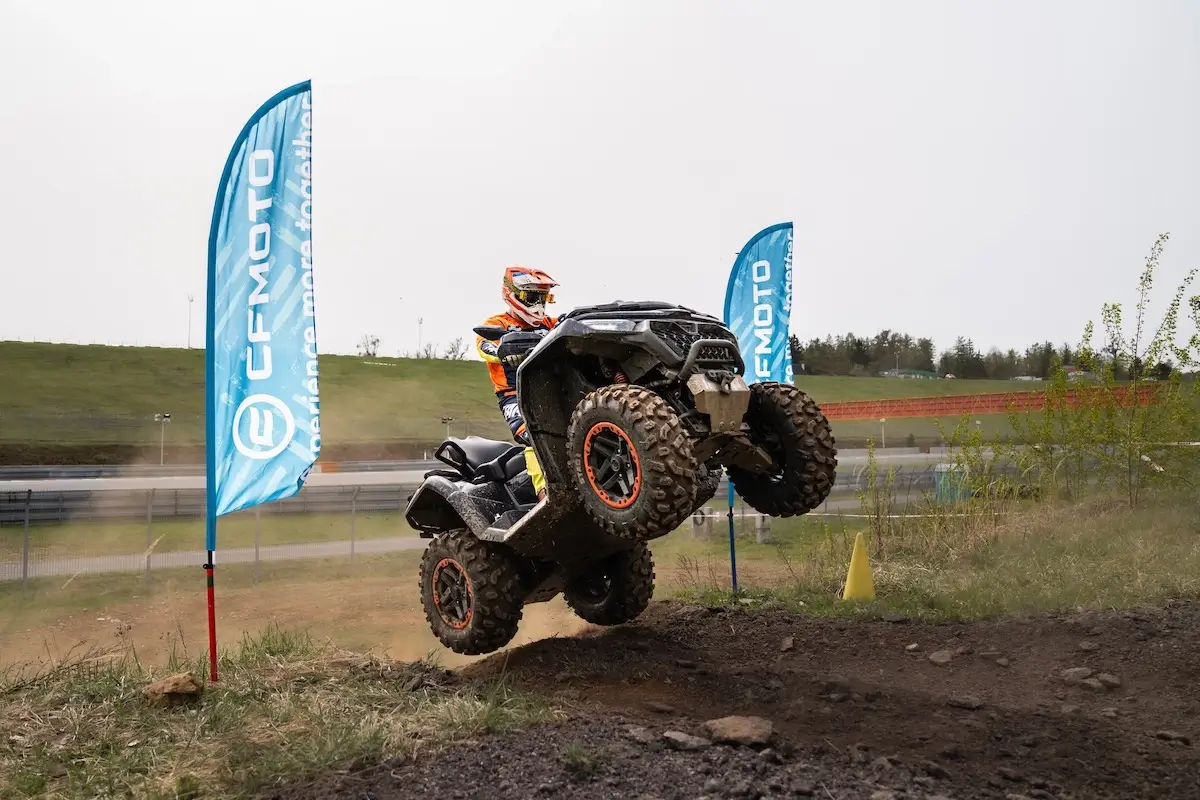 A person rides a CFMOTO cforce 1000 touring premium ATV, mid-air over a dirt mound on an off-road track, with two blue CFMOTO flags in the background.