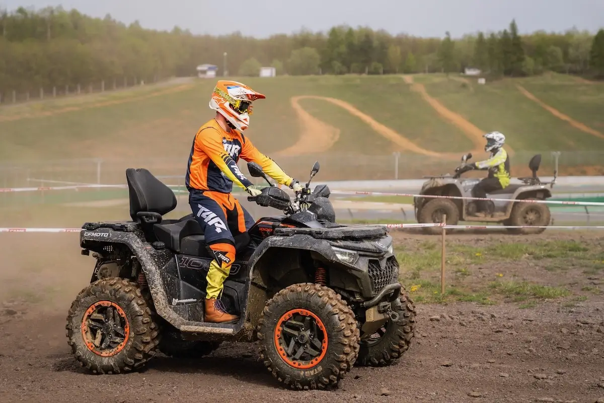 Two people ride ATVs on a dirt track outdoors; the closest rider, on a cforce 1000 touring premium, wears orange protective gear and helmet, while the other is farther in the background.