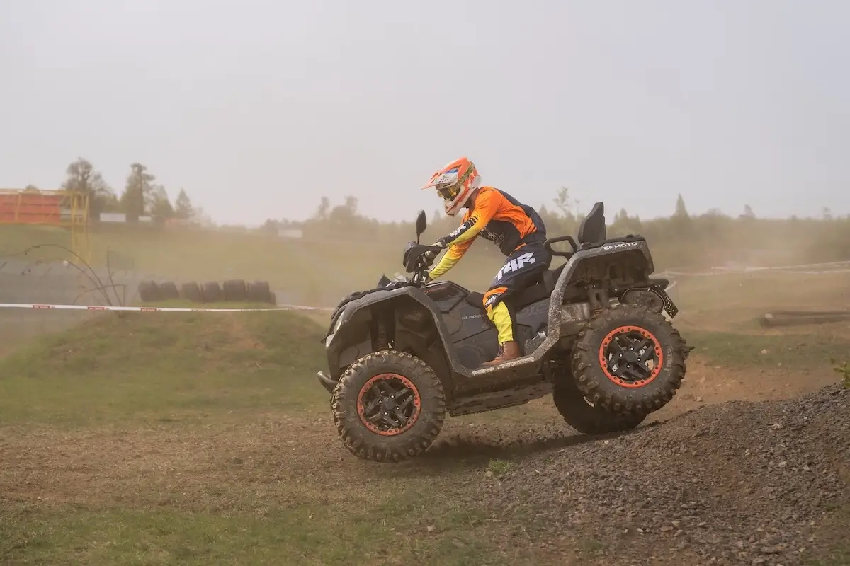 A person wearing orange and yellow motocross gear rides a cforce 1000 touring premium ATV over a dirt mound on an outdoor track.