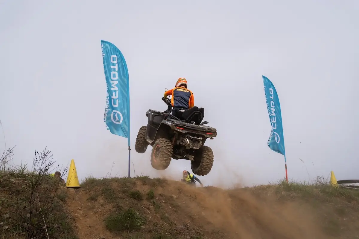 A person riding a CForce 1000 Touring Premium ATV is airborne over a dirt ramp between two blue flags with Cyrillic writing, surrounded by dust and grass.