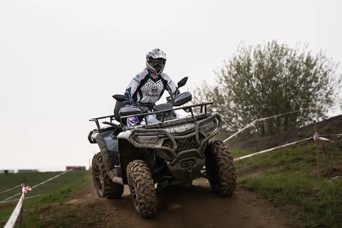 A person wearing a helmet and protective gear rides a CForce 850 Touring all-terrain vehicle on a dirt track, with grass and trees visible in the background.