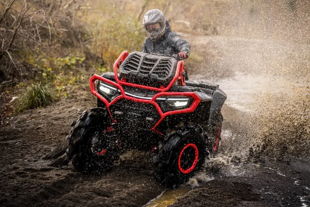 A person wearing a helmet rides a UFORCE U10 XL PRO ATV with red accents through mud and water, splashing both as they drive.