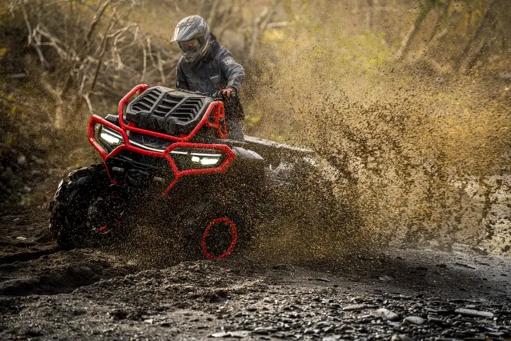 A person wearing a helmet rides a UFORCE U10 XL PRO ATV through muddy terrain, causing mud and water to splash up around the vehicle.