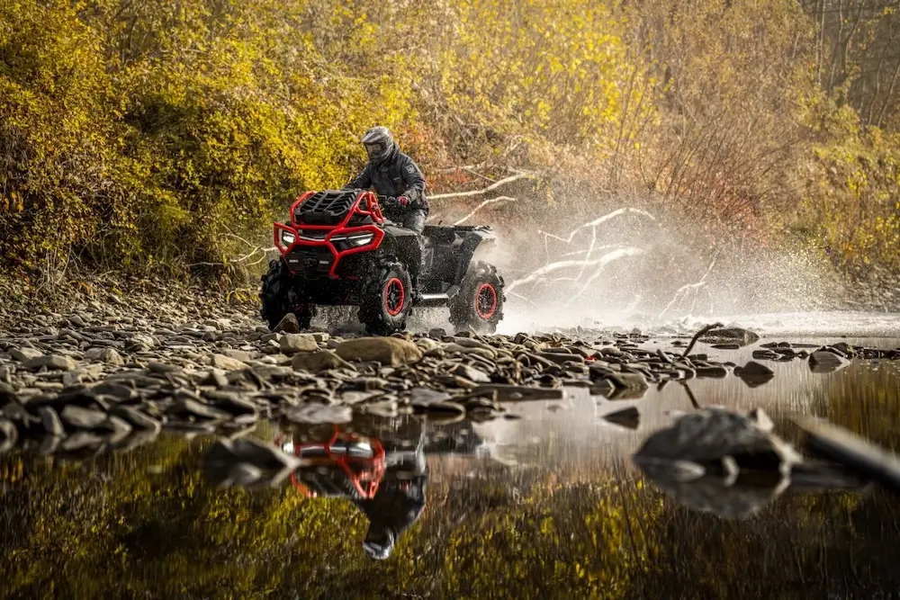 A person wearing a helmet rides a UFORCE U10 XL PRO ATV across a rocky riverside, with water splashing and trees with autumn foliage in the background.