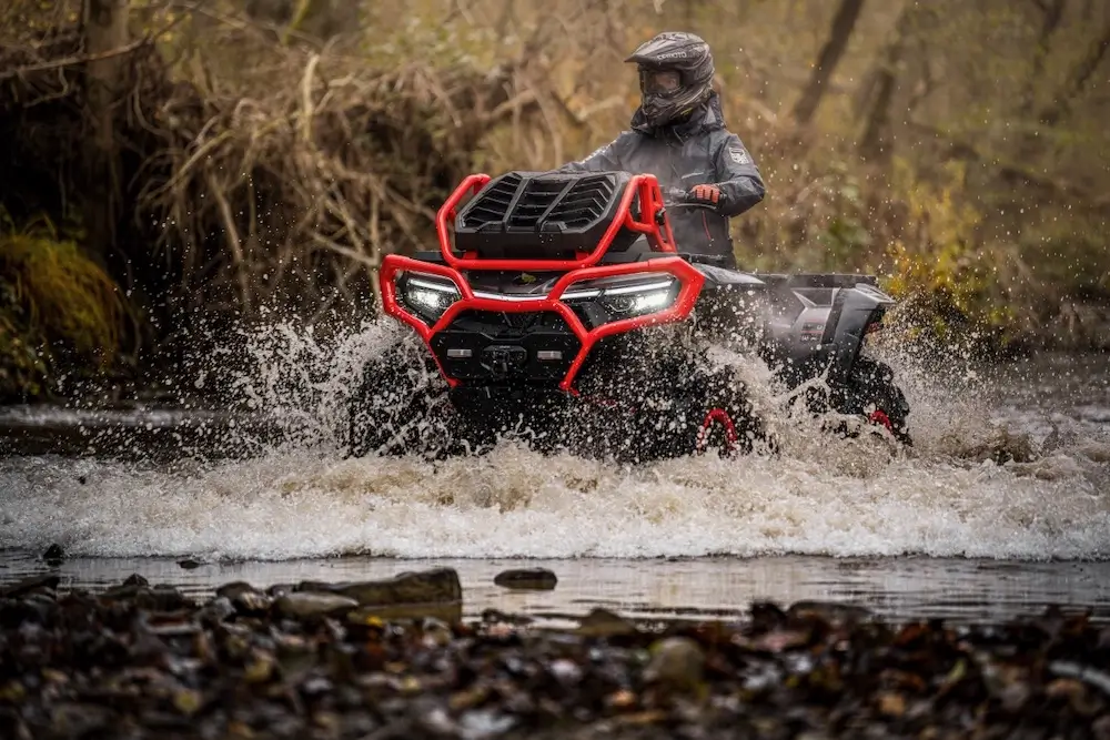 Wearing protective gear, a person rides a UFORCE U10 XL PRO ATV through a shallow, rocky creek, sending water splashing around the vehicle in a forested area.
