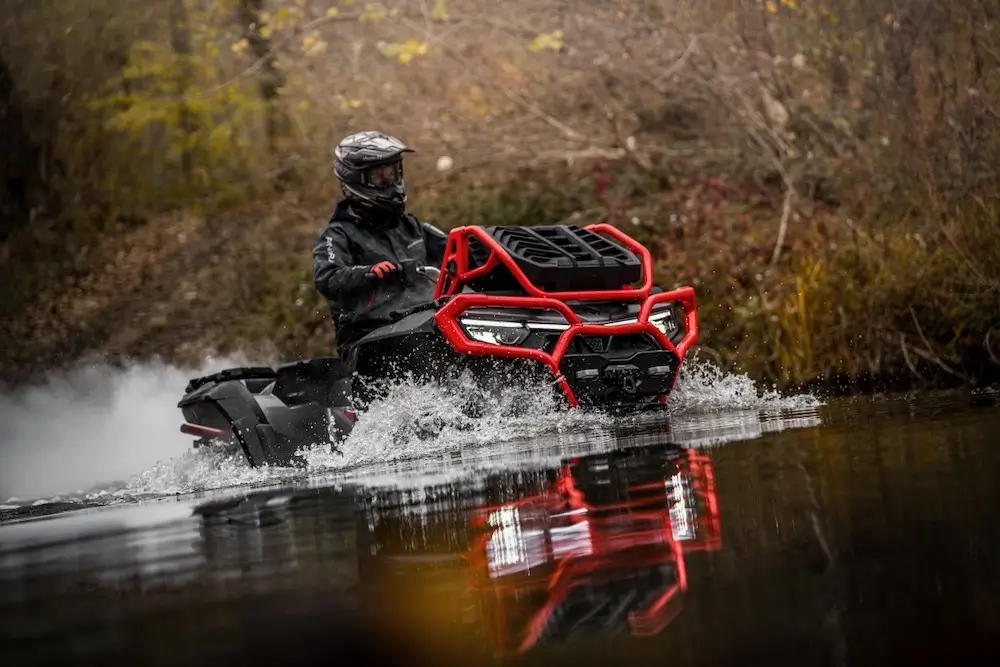 A person wearing a helmet and gear rides an ATV with a red grille, like the UFORCE U10 XL PRO, through shallow water in a wooded area.