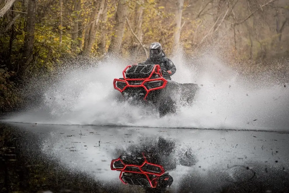 A person in full gear rides a UFORCE U10 XL PRO ATV through a shallow, wooded body of water, creating a large splash and reflection.
