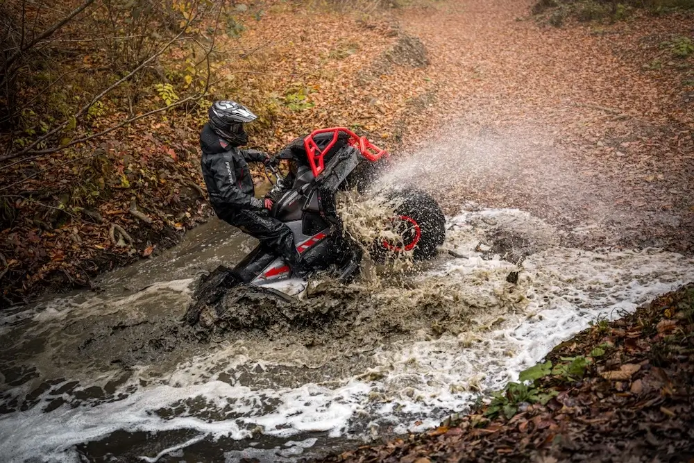 A person wearing protective gear rides a UFORCE U10 XL PRO ATV through a muddy stream in a forest with fallen leaves, causing water and mud to splash.