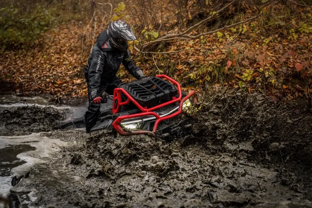 A person in protective gear stands next to a UFORCE U10 XL PRO all-terrain vehicle stuck in deep mud in a forested area.