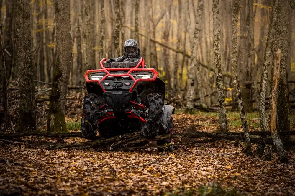 A person wearing a helmet rides a red UFORCE U10 XL PRO ATV through a forest with leaf-covered ground and bare trees.
