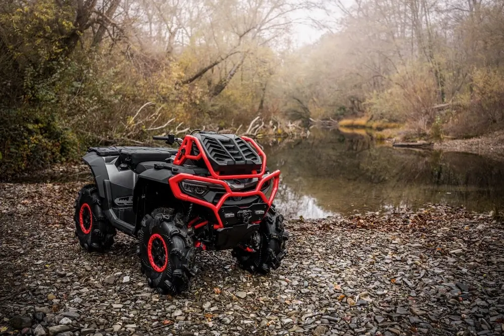 A black and red UFORCE U10 XL PRO all-terrain vehicle is parked on a rocky riverbank surrounded by leafless trees on a cloudy day.