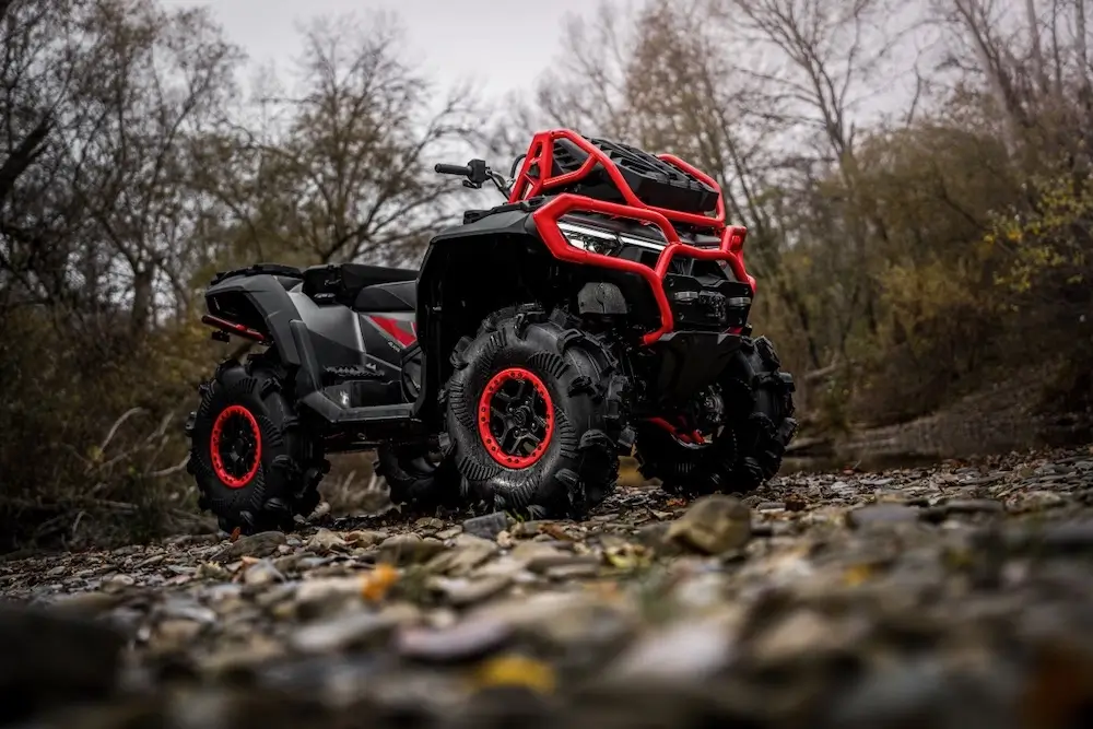 A black and red UFORCE U10 XL PRO all-terrain vehicle with large tires is parked on a rocky surface in a forested area under overcast skies.