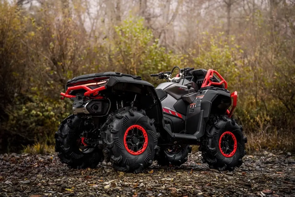 A black and red UFORCE U10 XL PRO all-terrain vehicle (ATV) with large, rugged tires is parked on rocky ground in front of dense, leafless trees and bushes.