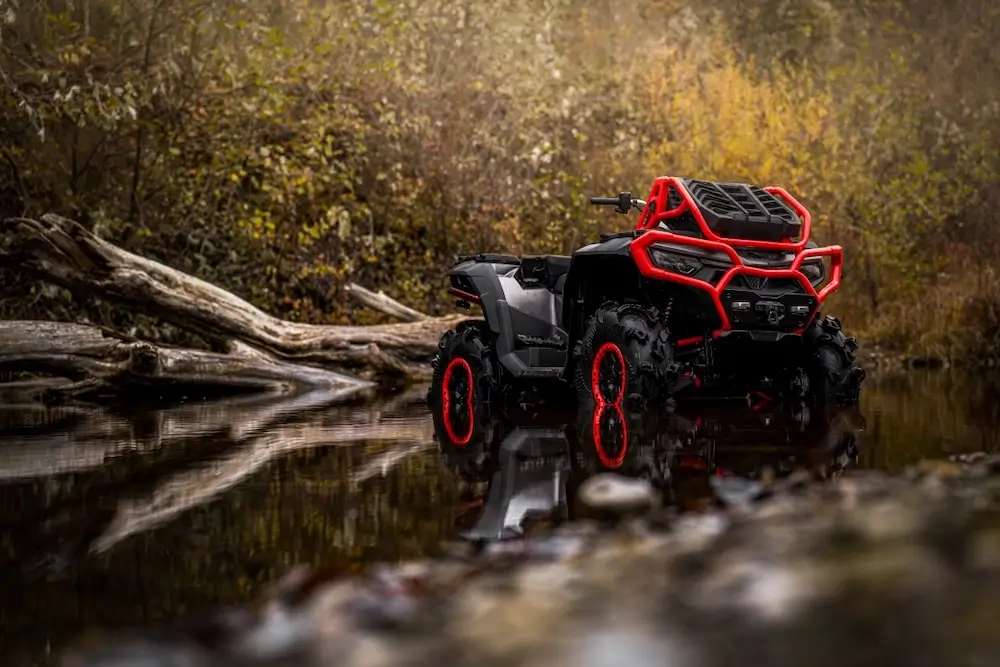 A black UFORCE U10 XL PRO ATV with red accents is parked in shallow water near fallen logs and dense foliage in a forest setting.