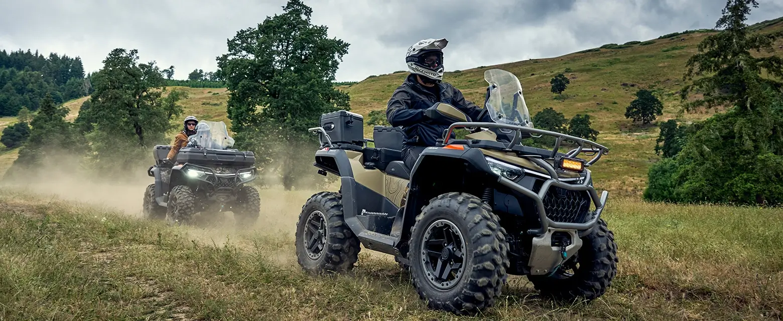 Two people riding CForce 1000 Touring Premium all-terrain vehicles on a dirt path, surrounded by trees and hills under a cloudy sky.