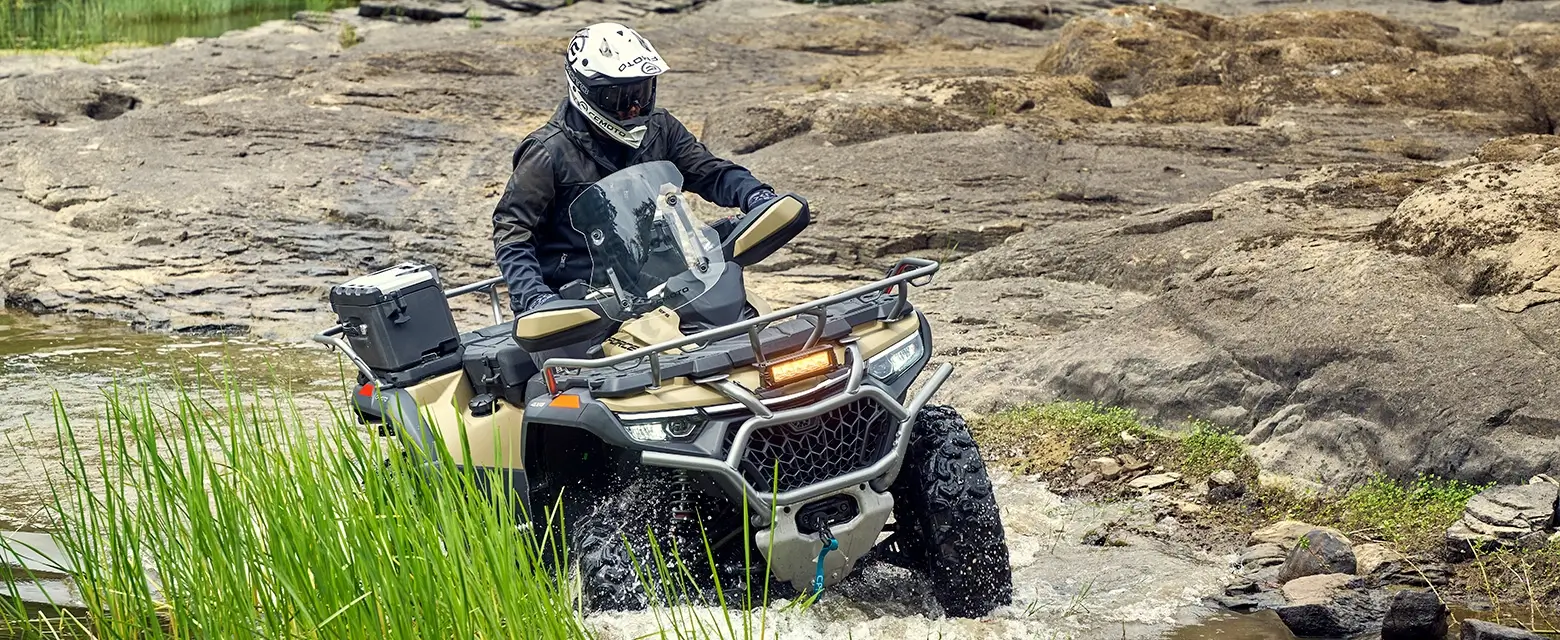 A person in protective gear expertly maneuvers a CForce 1000 Touring Premium ATV across the rugged terrain, navigating the rocky and shallow water area surrounded by lush grass and scattered stones.