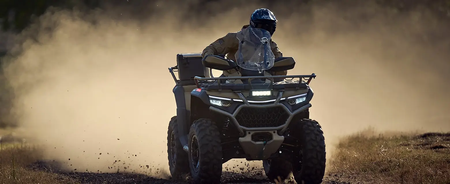 A person skillfully navigates a dirt path on their CFORCE 1000 Touring Premium ATV, dust swirling behind them. Clad in protective gear and a helmet, they embody both safety and excitement.
