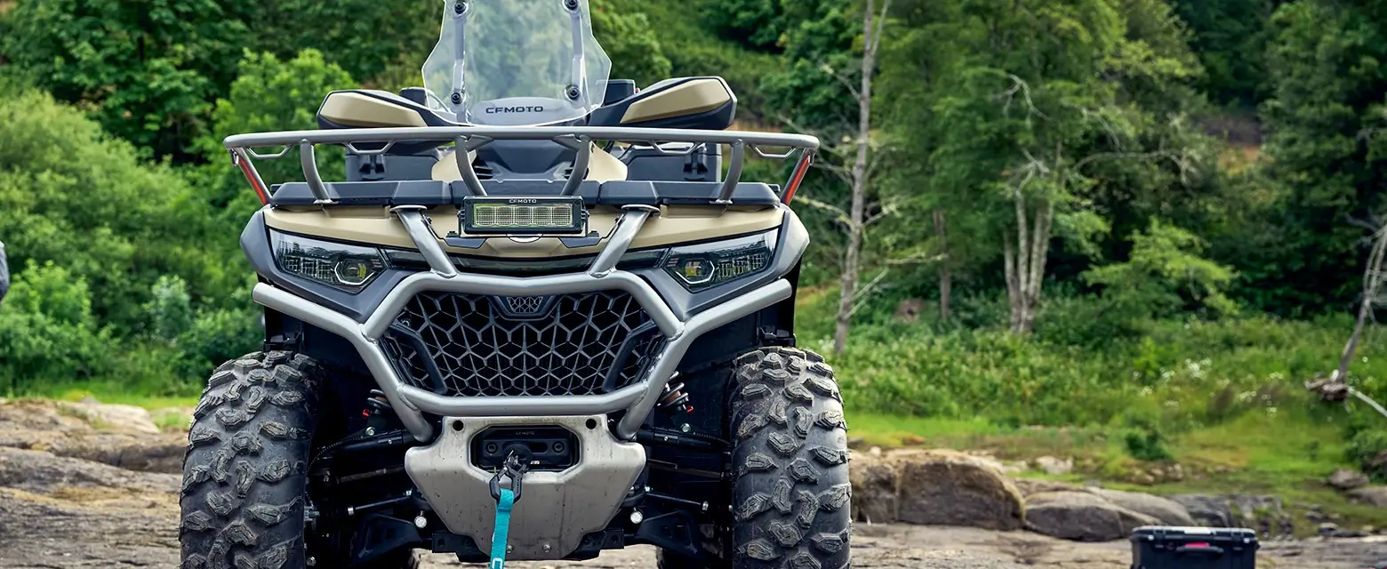 Front view of a CForce 1000 Touring Premium all-terrain vehicle with large tires parked on a rocky surface, surrounded by lush greenery.
