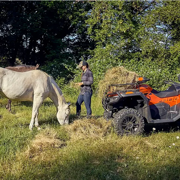 An equestrian stands near a white horse eating hay beside an orange ATV loaded with hay in a grassy field with trees in the background.