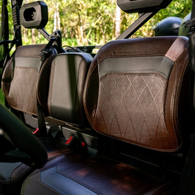 Comfort for 6 passengers Close-up of three brown and black leather seats with diamond stitching inside a 6-seater UTV, surrounded by lush greenery outside.