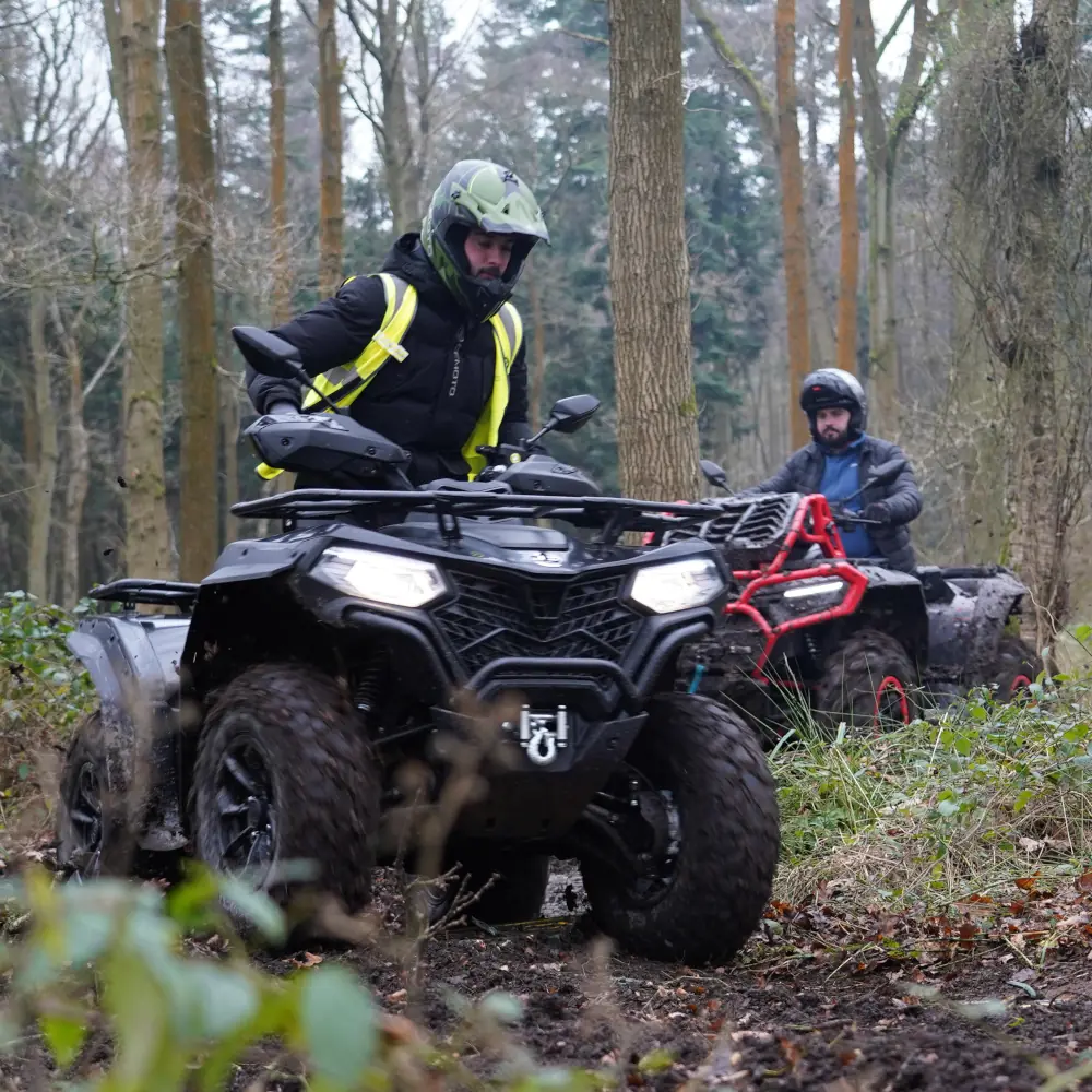 Two people ride a fleet of all-terrain vehicles through a muddy forest trail, wearing helmets and outdoor kit.