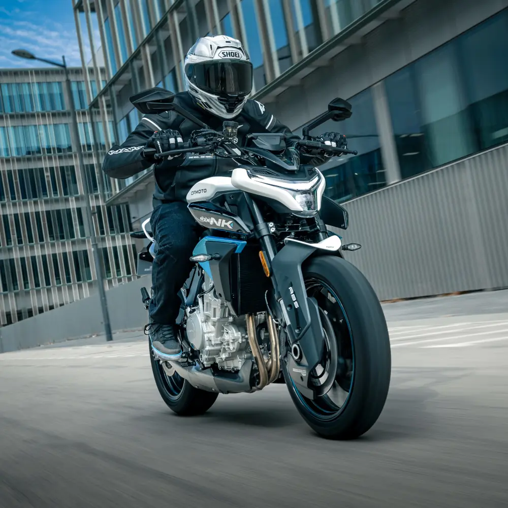 A motorcyclist wearing safety gear rides a blue and white commuter motorcycle on an empty urban street, with modern buildings in the background.
