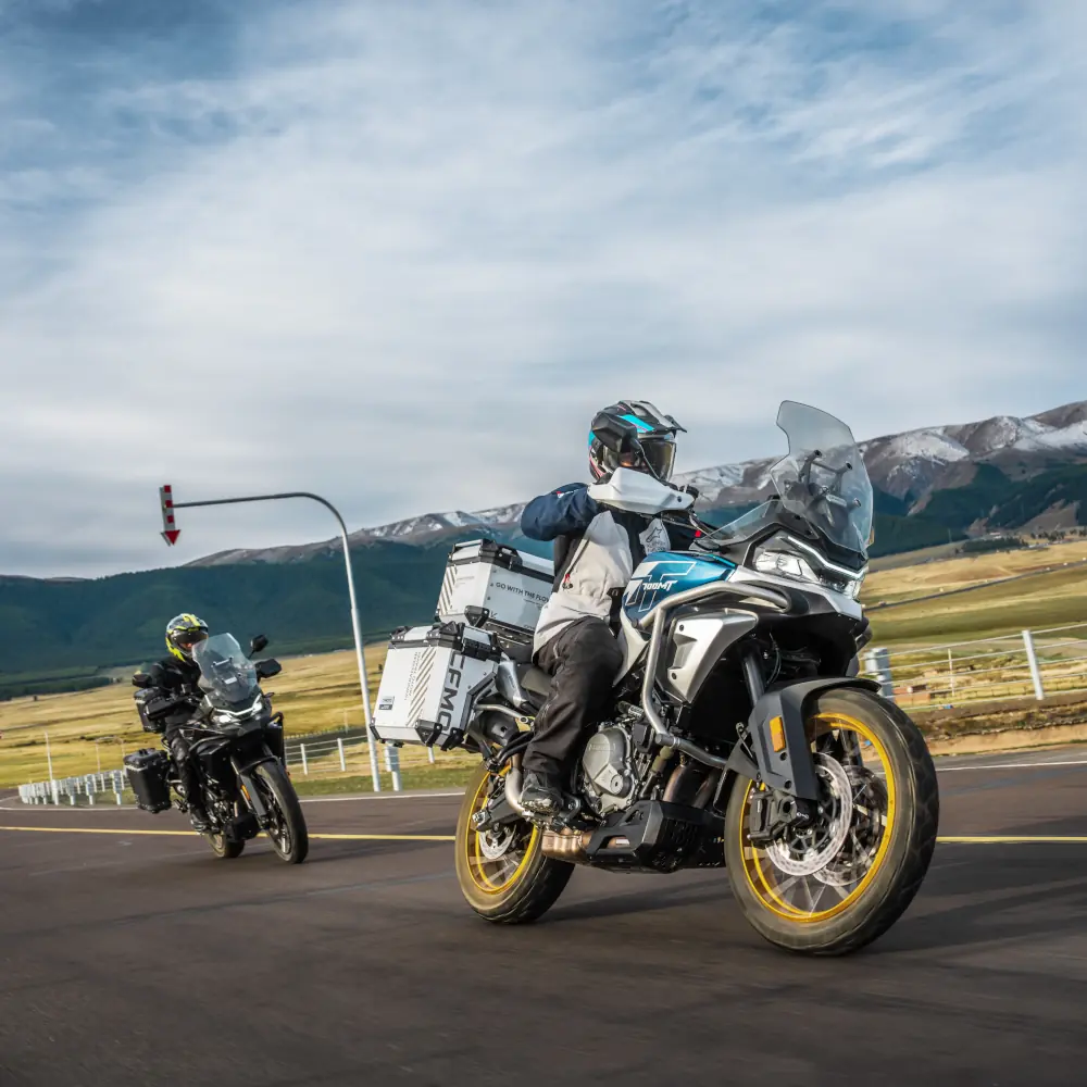 Two motorcyclists enjoy leisure riding on adventure touring motorcycles along a paved road, with mountains and fields stretching into the distance under a cloudy sky.