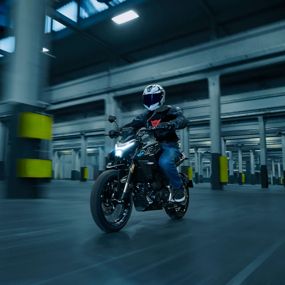 A person wearing a helmet and jacket rides a commuter motorcycle through a spacious, industrial-looking indoor area with columns.
