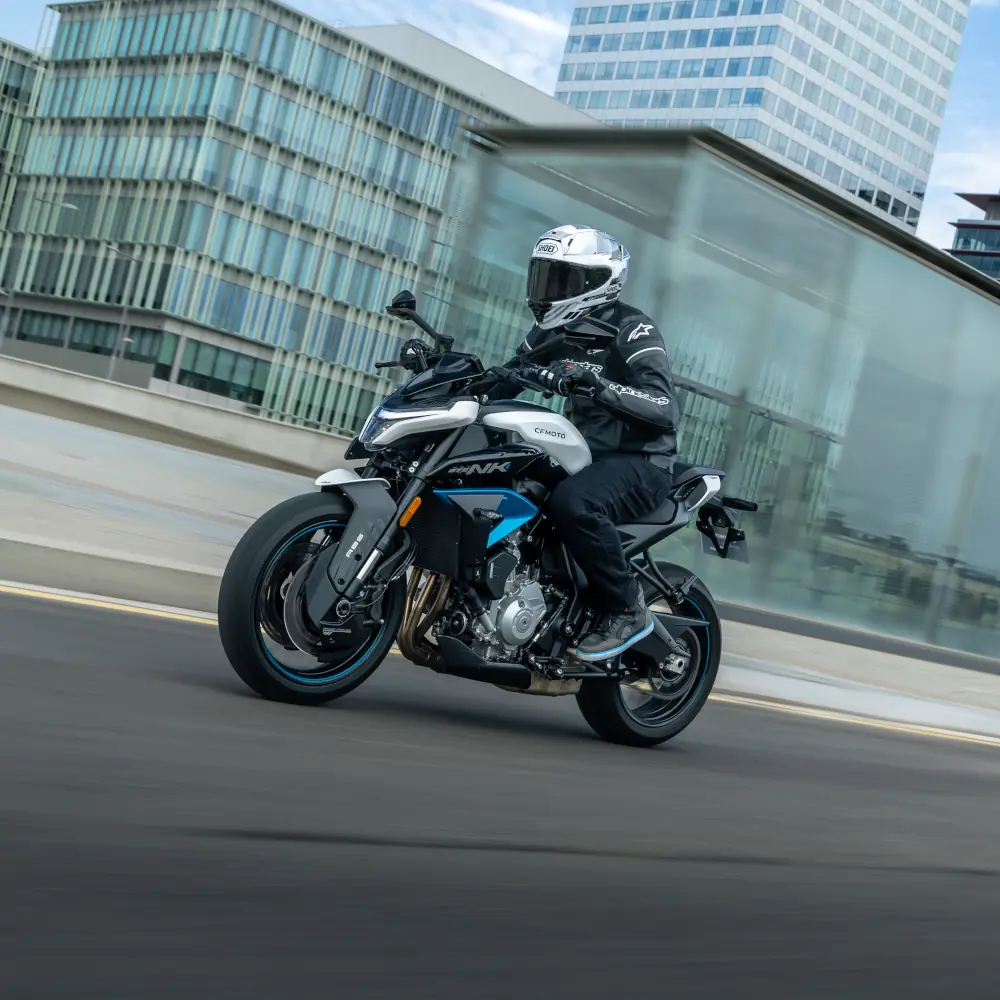 A motorcyclist in full riding gear enjoys leisure riding on a white and blue sport motorcycle through an urban road, framed by modern glass buildings in the background.