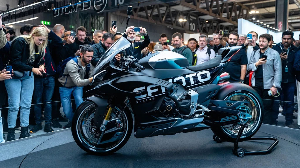 A crowd gathers around and photographs a black and white FIOTO racing motorcycle displayed on a stand at an indoor event, alongside the innovative CFMOTO CFORCE TECH GEN 4 Concept.