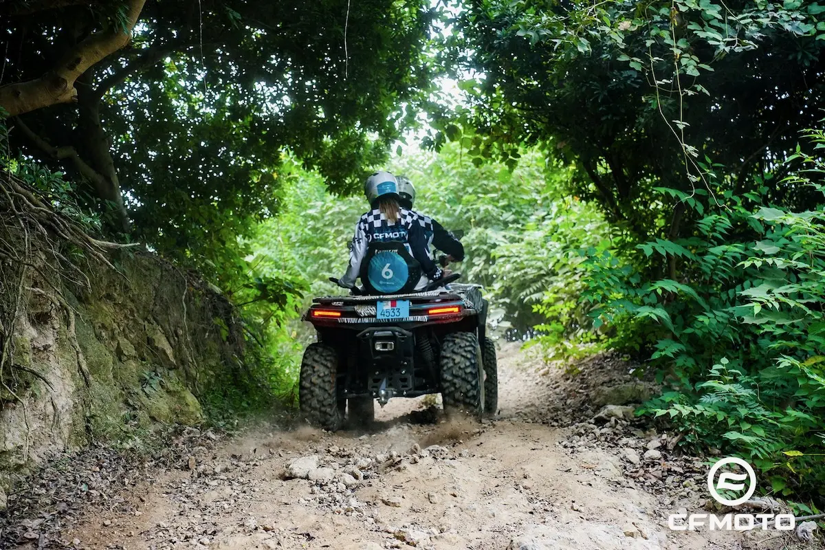 Two people ride an ATV on a rocky, dirt trail surrounded by dense green foliage, demonstrating ATV safety. The vehicle and riders' gear feature the ECFMOTO logo.