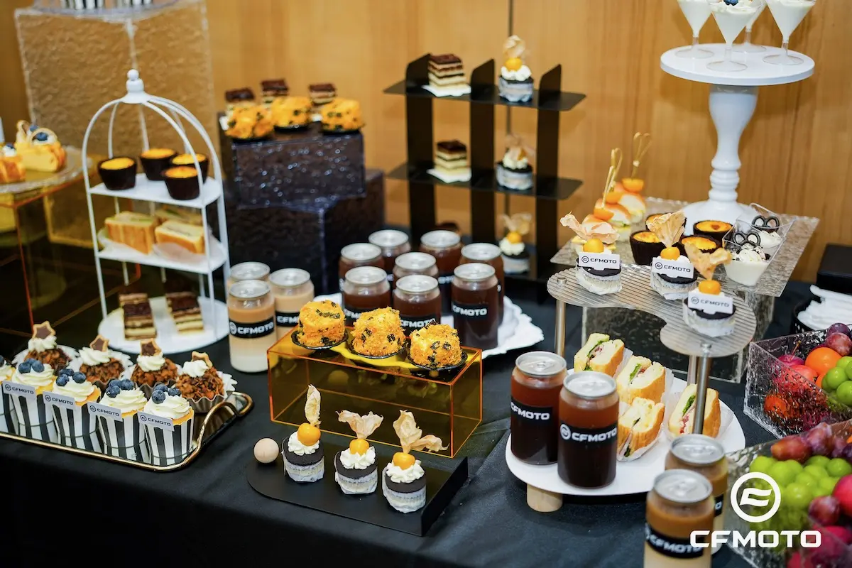 A dessert table displays cakes, pastries, fruit, sandwiches, and drinks in jars labeled "CFMOTO," arranged neatly on stands and trays, celebrating ATV safety with style and attention to detail.