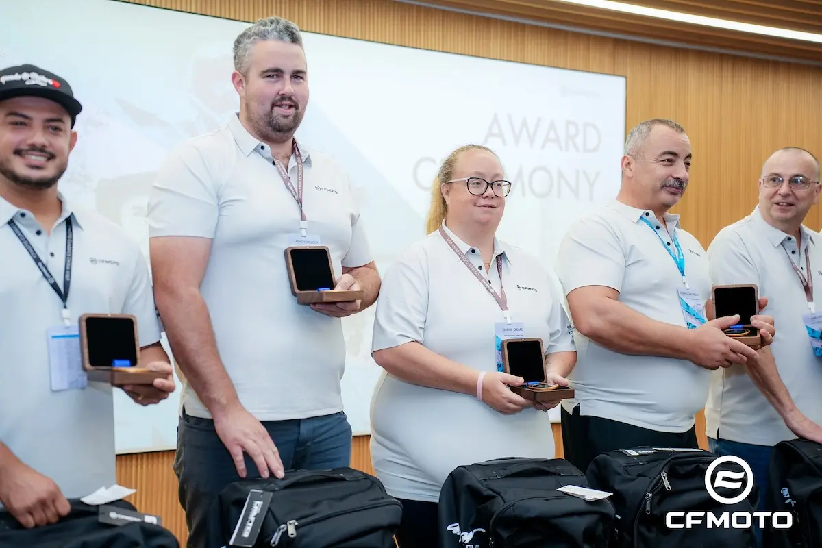 Five people in white shirts stand in a row holding award boxes at a ceremony, highlighting ATV safety, with backpacks and a CFMOTO logo visible in the foreground.