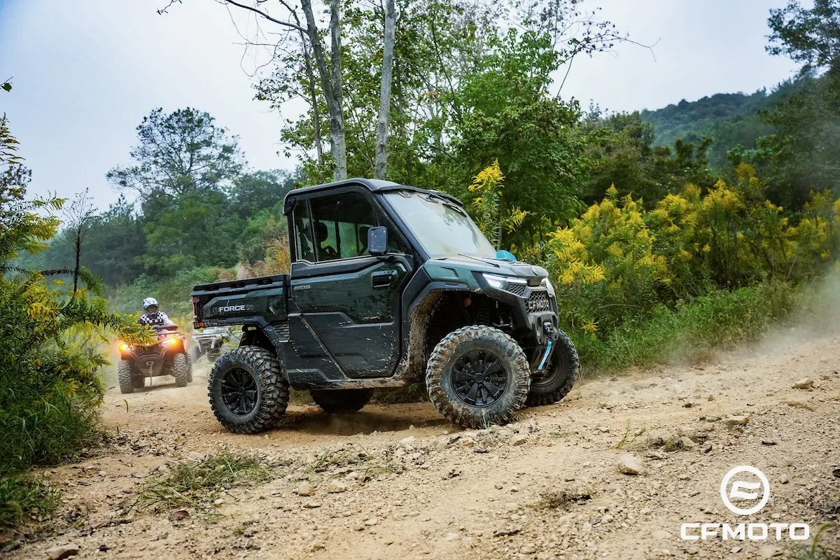 A black CFMOTO utility vehicle drives on a rocky dirt trail through a forested area, with a focus on ATV safety as a rider follows behind on an ATV. The CFMOTO logo is visible in the bottom right corner.