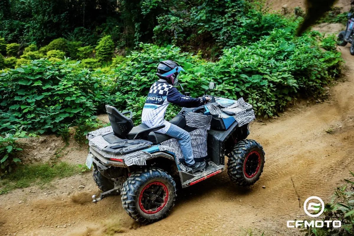 Riding a CFMOTO all-terrain vehicle on a dirt trail surrounded by greenery, this person follows ATV safety guidelines, wearing protective gear and a helmet.