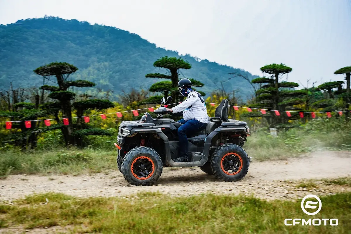 A person demonstrates ATV safety by wearing a helmet while riding an ATV on a dirt path lined with trimmed trees and mountains in the background; the CFMOTO logo is visible.