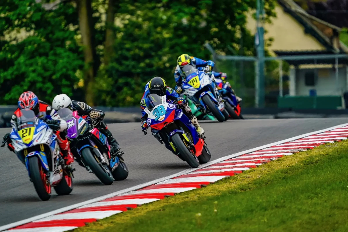 _DSC0279 Four motorcycle racers from Moto Twelve lean into a bend at Oulton Park racetrack, closely following each other, with trees and buildings in the background.