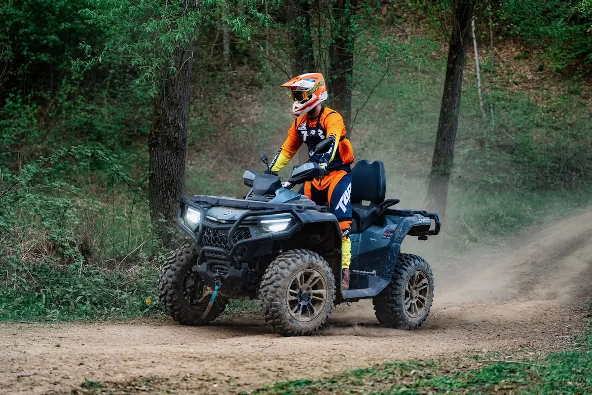 A person wearing orange motocross gear and helmet rides a black CForce 850 Touring ATV on a dirt path through a forested area.