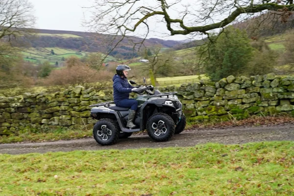 A person wearing a helmet and outdoor clothing rides an estate management utility vehicle on a dirt path next to a stone wall in a rural, hilly landscape.