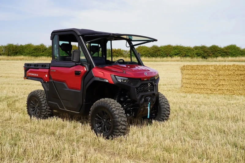 A red off-road utility vehicle with a black roof is parked on a harvested field beside stacked hay bales, hinting at the scenic motorcycle routes that wind through the trees in the background.