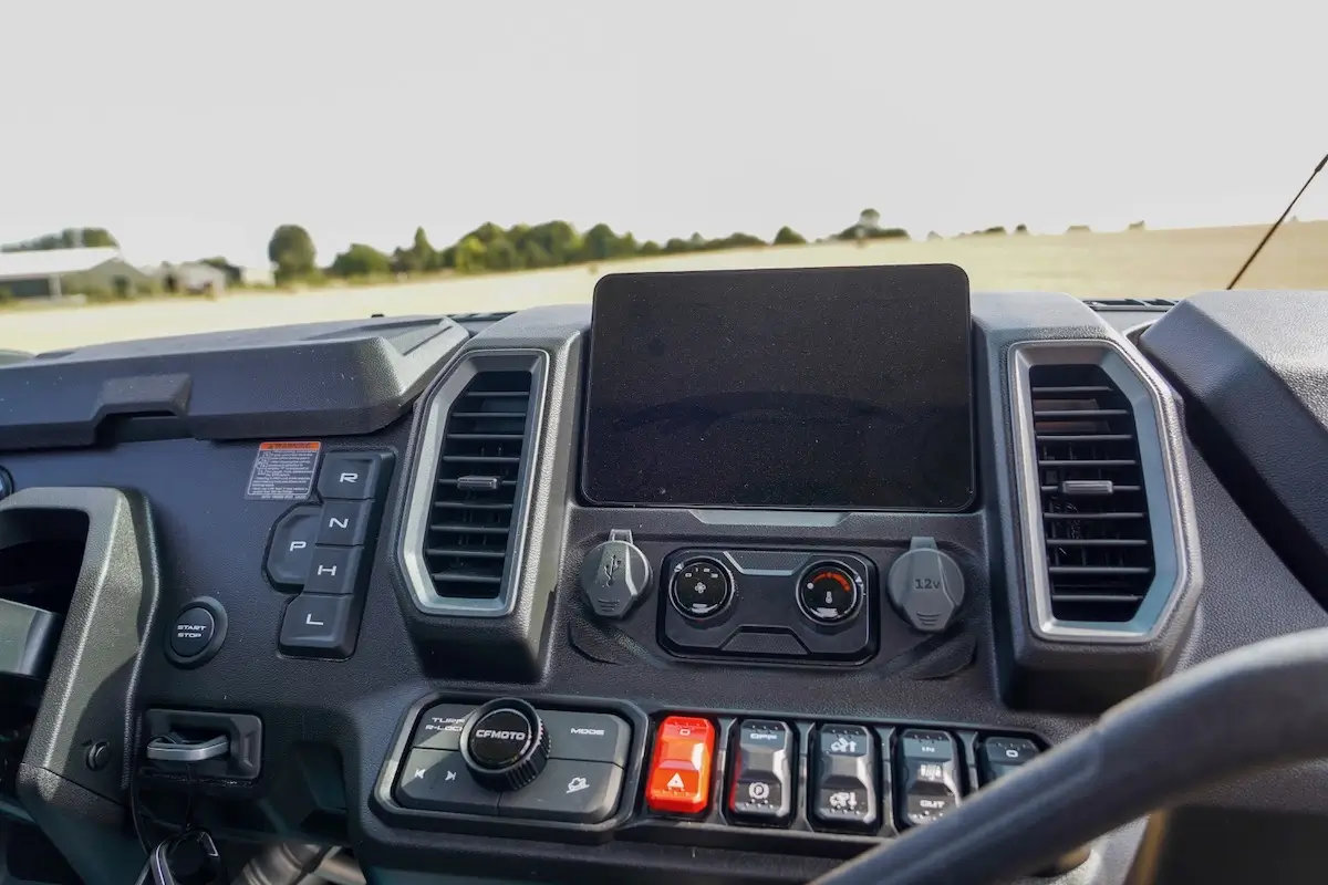 Dashboard of the UFORCE U10 PRO HIGHLAND featuring a central touchscreen, control buttons, air vents, gear selector, and climate controls, with a blurred outdoor landscape visible through the windshield.