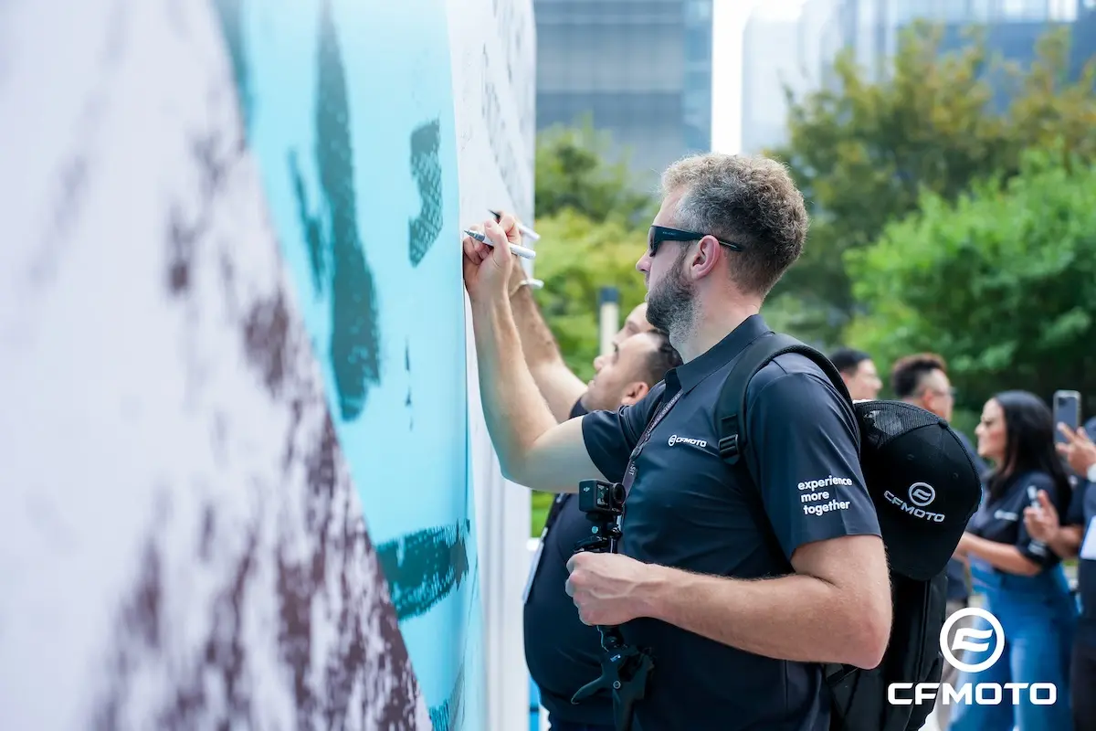 A man wearing a CFMOTO shirt and backpack writes about ATV safety on a large outdoor mural, surrounded by others also participating in the activity.